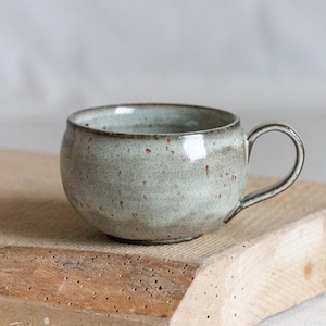 Könnte beinhalten: Eine hellgrüne Keramiktasse mit gesprenkeltem Glase und runder Form. Die Tasse hat einen braunen Rand und einen Henkel.