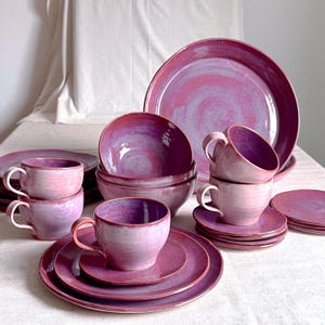 Op de afbeelding: Een verzameling handgemaakt keramisch servies in tinten paars en roze. De set bevat kopjes, schotels, kommen en borden van verschillende formaten. De stukken hebben een glanzende afwerking en een uniek, wervelend kleurenpatroon.