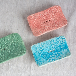 Può includere: Tre portasapone in ceramica rosa, turchese e verde. Ogni piatto rettangolare presenta un motivo floreale in pizzo e due fori di drenaggio. I piatti sono disposti su una superficie di lino bianco.