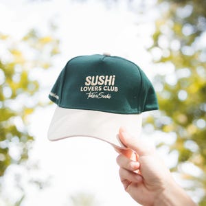 Puede incluir: Una gorra de béisbol verde y blanca con el texto "Sushi Lovers Club" y "Take Sushi" bordado en la parte delantera.