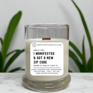 May include: A clear glass candle with a wooden wick and a white label. The label reads "I MANIFESTED & GOT A NEW ZIP CODE." The candle is filled with a creamy wax and is labeled as 8oz, hand-poured in Michigan.