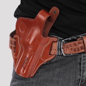 Könnte beinhalten: Ein braunes Lederholster mit einem genähten Design und dem Wort "Marshal" geprägt auf der Seite. Das Holster ist an einem braunen Gürtel mit Krokodilmuster befestigt.