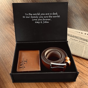 May include: A black gift box with a brown leather wallet and belt. The wallet has the initials "TM" and the name "THOMAS MOORE". The belt has a silver buckle. The box lid has the text: "To the world you are a dad, to our family you are the world. Love you forever, Amy & John."