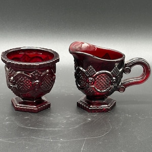 May include: Two pieces of ruby red glass tableware. One is a small, open bowl with a decorative pattern. The other is a creamer with a handle and spout, also with a decorative pattern. Both pieces have a hexagonal base.