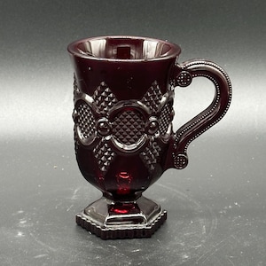 May include: A ruby red glass mug with a handle and a decorative pattern. The mug has a pedestal base and a detailed design around the body. The handle is curved and has a small decorative element at the top. The mug is likely vintage or antique.