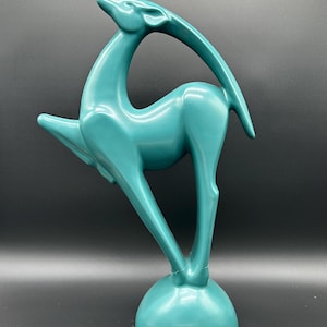 May include: A teal ceramic sculpture of a gazelle in a dynamic pose. The gazelle has a slender body, curved horns, and is mounted on a round base. The sculpture is a solid color and has a glossy finish. The artwork is a decorative item.