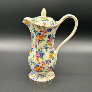 May include: A vintage floral-patterned ceramic pitcher with a lid. The pitcher features a cream-colored base with a colorful floral design in pink, blue, yellow, and orange. It has a curved handle and a spout for pouring. The lid is topped with a small finial.