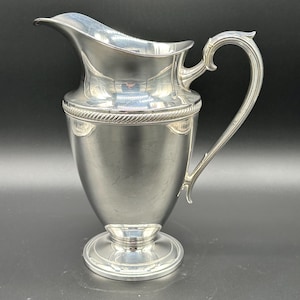May include: A silver-colored pitcher with a curved spout and ornate handle. The pitcher has a round base and a decorative rope-like detail around the top. The pitcher is made of a shiny metal and reflects the surrounding environment.
