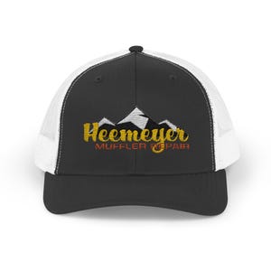 May include: Black and white trucker hat with the text "Heemeyer Muffler Repair" in gold and orange. The hat features a mountain graphic above the text. The hat has a black brim and crown with a white mesh back.