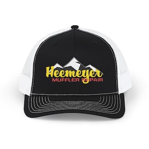 May include: Black and white trucker hat with the text "Heemeyer Muffler Repair" in yellow and red. The hat features a mountain graphic above the text. The hat has a curved brim and a mesh back.