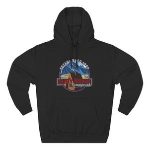 Può includere: Felpa con cappuccio nera con grafica di un paesaggio montano. Il design include una baita e una catena montuosa in un emblema circolare, con la scritta "Ruby Ridge Campground" e "Established 1992".