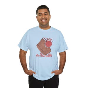 May include: Light blue t-shirt with a graphic design. The design features a brown wooden block with a red and silver circular object on top, and the words "Livin' on the edge" in red script.