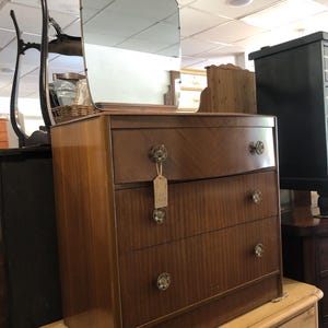 May include: A vintage wooden chest of drawers with a mirror. The chest has three drawers with decorative knobs and a curved front. The mirror is arched at the top. The wood has a warm, reddish-brown tone.