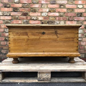May include: An antique wooden chest in light brown. The chest is rectangular with a hinged lid and sits on four rounded feet. The background is a brick wall. The chest is likely made of pine.