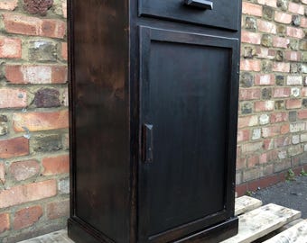 Puede incluir: Un mueble de madera teñida oscura con un cajón y una puerta. El mueble es rectangular con un diseño sencillo. El cajón tiene un pequeño tirador rectangular. La puerta tiene un tirador vertical. El mueble está sobre un palet de madera.