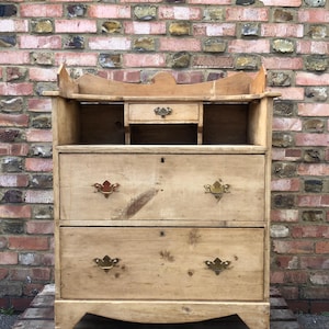 May include: A light-coloured wooden chest of drawers with two large drawers and a small top drawer. It features decorative brass handles and a scalloped top. The wood grain is visible, and the piece appears to be antique.