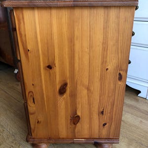 K&ouml;nnte beinhalten: Eine rechteckige Seitenwand eines Holzschranks mit einer nat&uuml;rlichen hellbraunen Oberfl&auml;che, sichtbarer Holzmaserung und dunklen, kreisf&ouml;rmigen Astl&ouml;chern. Das M&ouml;belst&uuml;ck hat eine flache Oberkante und steht auf runden, kugelf&ouml;rmigen F&uuml;&szlig;en.