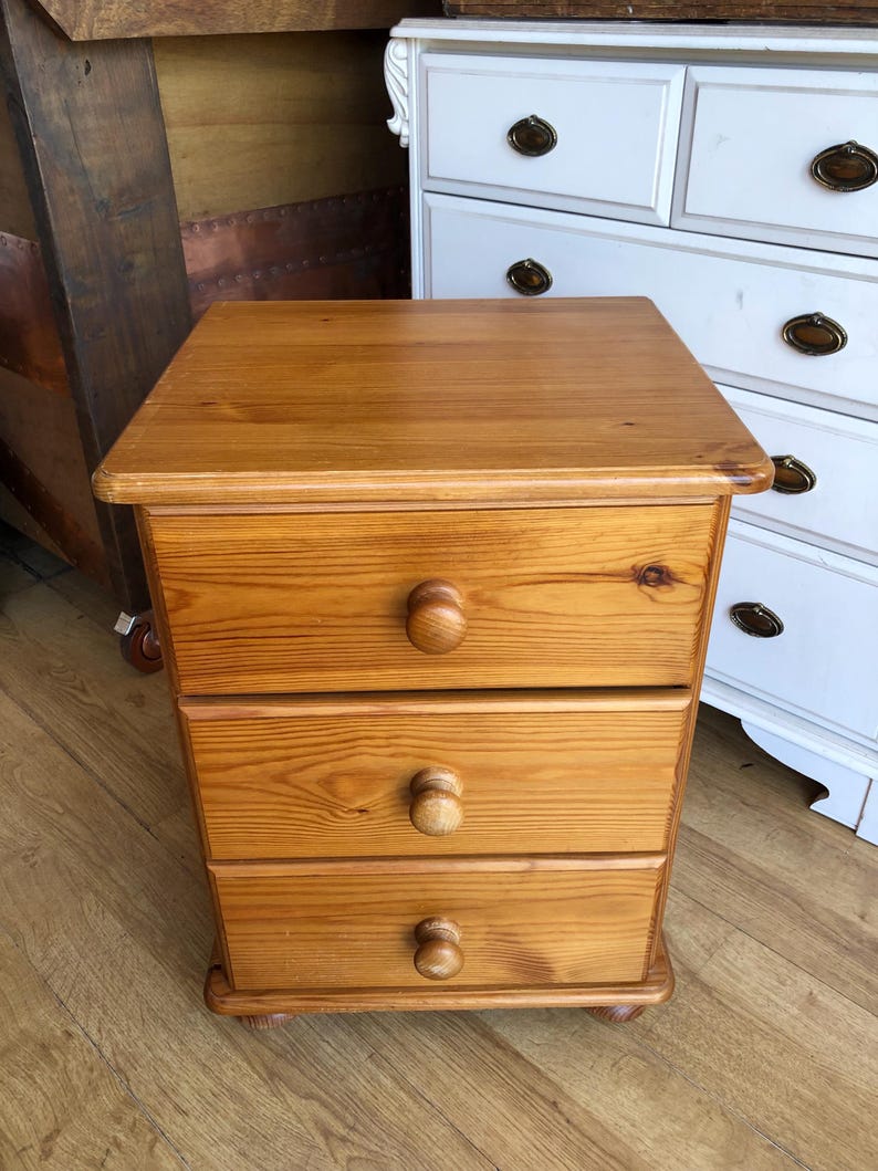 K&ouml;nnte beinhalten: Ein rechteckiger Nachttisch aus Holz mit einer nat&uuml;rlichen, hellbraunen Holzmaserung. Das M&ouml;belst&uuml;ck verf&uuml;gt &uuml;ber drei &uuml;bereinander angeordnete Schubladen, die jeweils mit einem runden Holzknauf ausgestattet sind. Es steht auf vier kleinen F&uuml;&szlig;en.