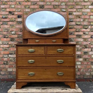 May include: A classic wooden dressing table with an oval mirror. The chest of drawers features brass handles and sits on a wooden pallet. The wood has a warm, reddish-brown tone.