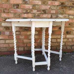 May include: A white, antique drop-leaf table with a round top and four spiral-turned legs. The table is painted white and has a distressed finish. The table is set against a brick wall. The table's height is approximately 76 cm.