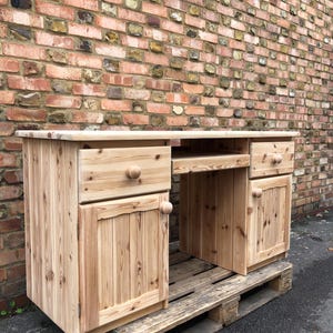 May include: A light-coloured wooden desk with two drawers and two cabinet doors. The desk has a cutout for a chair and is sitting on a wooden pallet. The background is a brick wall.