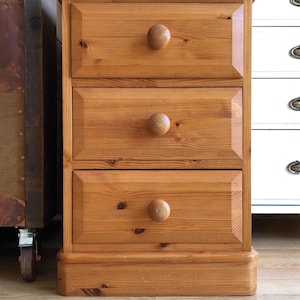 May include: A light brown wooden bedside table featuring three stacked drawers. Each drawer is equipped with a single round wooden knob and has a beveled rectangular frame design. The wood grain is visible throughout the entire structure.