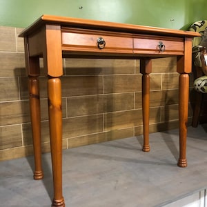May include: A wooden desk with two drawers and ring pulls. The desk has four rounded legs and a rectangular top. The wood has a warm, reddish-brown tone. The desk is approximately 76 cm wide and 51 cm deep.