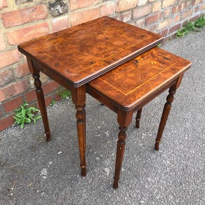 May include: A pair of antique nesting tables. The tables feature a rectangular top with a dark wood grain and a lighter border. The legs are turned and tapered. The larger table is approximately 61 cm wide and 46 cm deep.
