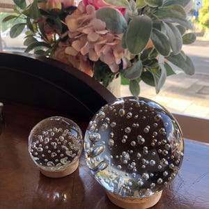 May include: Two clear glass spheres with internal air bubbles, sitting on small wooden bases. The spheres are different sizes, with the larger one in the foreground. A bouquet of pink and white flowers is in the background.
