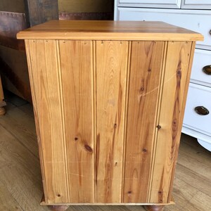 K&ouml;nnte beinhalten: Ein rechteckiger Holzschrank mit hellbrauner Oberfl&auml;che, bei der die Maserung und Astl&ouml;cher des Holzes sichtbar sind. Die Front besteht aus vertikalen Paneelen im Nut-und-Feder-Design und ruht auf vier kleinen, runden Holzf&uuml;&szlig;en.
