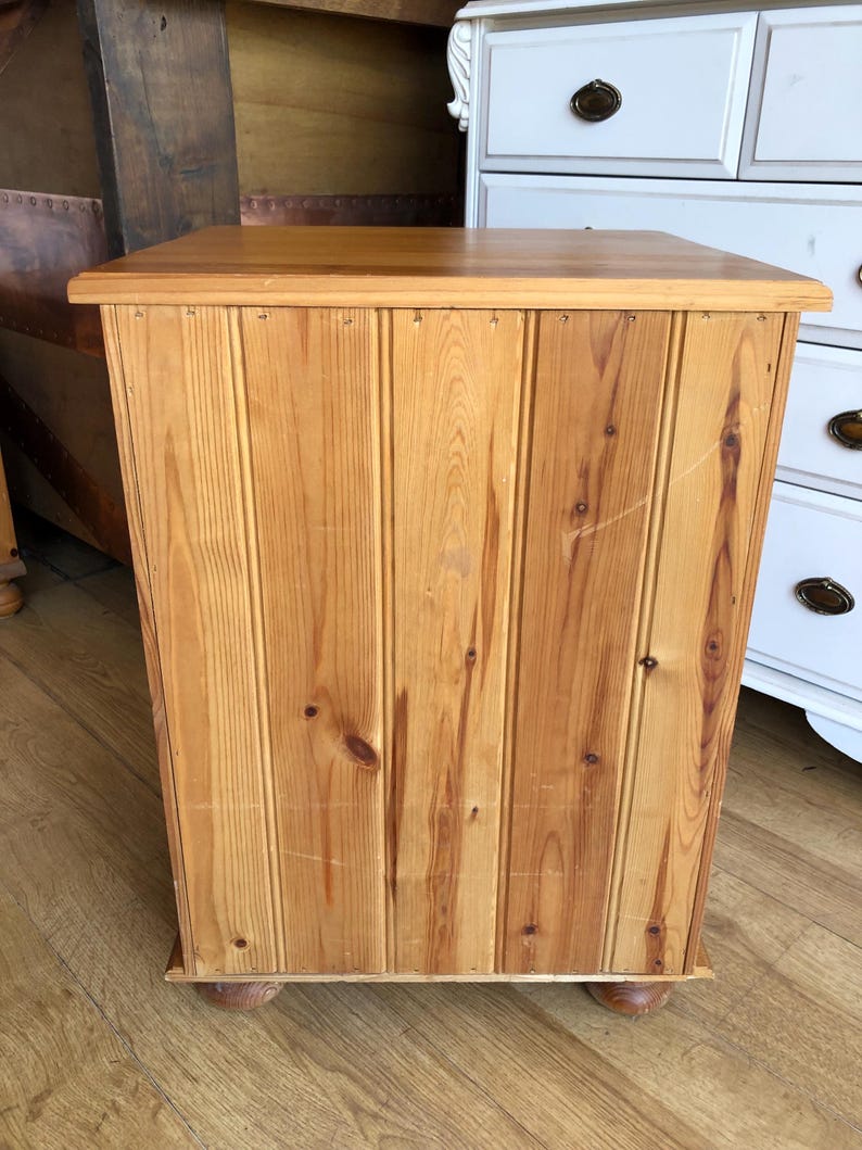 K&ouml;nnte beinhalten: Ein rechteckiger Holzschrank mit hellbrauner Oberfl&auml;che, bei der die Maserung und Astl&ouml;cher des Holzes sichtbar sind. Die Front besteht aus vertikalen Paneelen im Nut-und-Feder-Design und ruht auf vier kleinen, runden Holzf&uuml;&szlig;en.