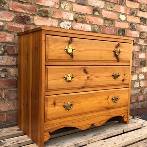 May include: A light brown wooden chest of drawers featuring three horizontal drawers with visible wood grain and knots. Each drawer is equipped with two ornate brass swing handles. The base of the chest has a decorative scalloped apron design.