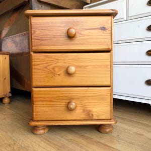 May include: A light brown wooden bedside cabinet featuring three stacked drawers, each fitted with a single round wooden knob. The piece is supported by four rounded bun feet and displays a visible natural wood grain pattern throughout its surface.