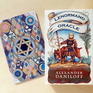 Puede incluir: Una baraja Lenormand Oracle de Alexander Daniloff con la ilustración de una persona con un perro y un estandarte, junto a una carta con un patrón geométrico de diamantes y una estrella de seis puntas en tonos azules, rosas y dorados.