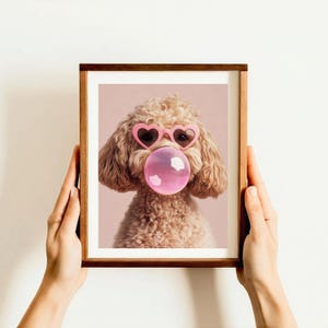 May include: A framed print of a light brown poodle wearing pink heart-shaped sunglasses and blowing a pink bubble. The background is a light pink color.