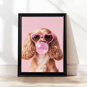 Puede incluir: Impresión enmarcada de un Cocker Spaniel con gafas de sol rosas en forma de corazón y soplando una burbuja de chicle. El perro tiene pelo ondulado de color marrón claro, sobre un fondo rosa. El marco es negro.