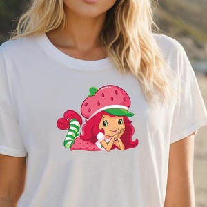 May include: White t-shirt featuring a colorful graphic of a cartoon character. The character has red hair, a strawberry hat, and a green and white striped tail. The character is lying down with a smile.