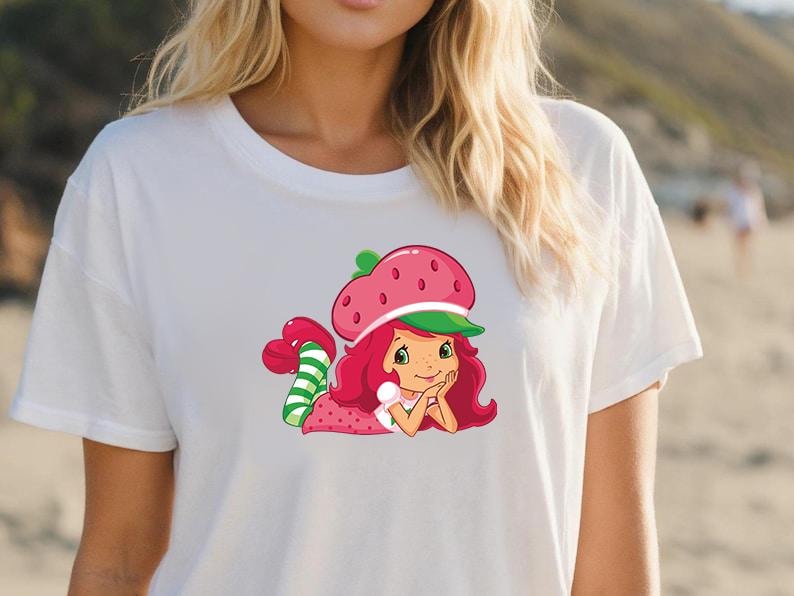 May include: White t-shirt featuring a colorful graphic of a cartoon character. The character has red hair, a strawberry hat, and a green and white striped tail. The character is lying down with a smile.