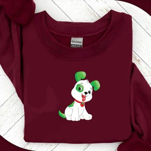 May include: A maroon sweatshirt featuring a cartoon dog graphic. The dog is white with green accents on its ears and spots, a red collar, and a smiling expression. The sweatshirt is made of a heavy blend fabric.