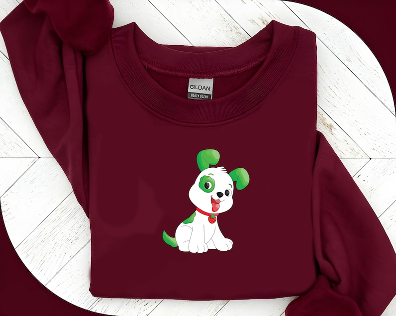 May include: A maroon sweatshirt featuring a cartoon dog graphic. The dog is white with green accents on its ears and spots, a red collar, and a smiling expression. The sweatshirt is made of a heavy blend fabric.