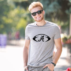 Pu&ograve; includere: Una maglietta grigia con un disegno di football americano nero, la lettera "A" e il testo "Roll Tide". La maglietta &egrave; realizzata in un materiale morbido ed &egrave; adatta per l'abbigliamento casual.