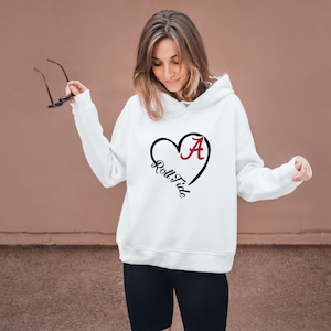 Pu&ograve; includere: Felpa con cappuccio bianca con grafica a cuore nero contenente la lettera "A" bordeaux e le parole "Roll Tide" in corsivo nero. La felpa ha un cappuccio con coulisse e maniche lunghe. La persona indossa pantaloncini neri.