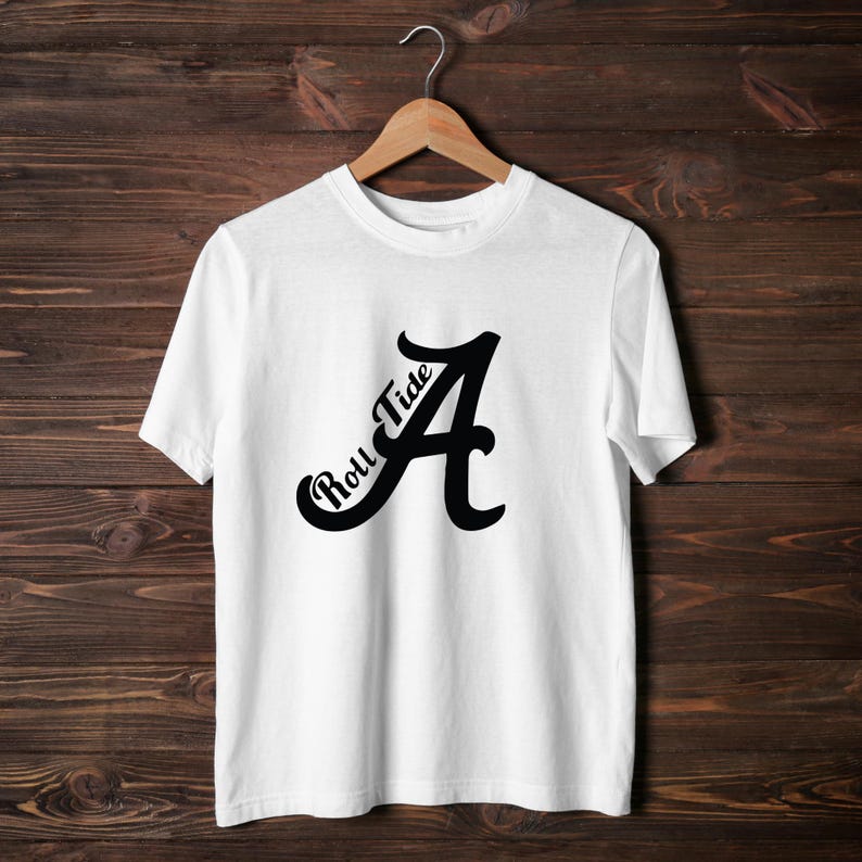 Pu&ograve; includere: T-shirt bianca con grafica nera della lettera "A" e la scritta "Roll Tide" in corsivo. La maglietta &egrave; appesa a una gruccia di legno su uno sfondo di legno scuro.
