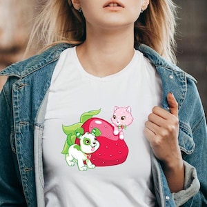 May include: White t-shirt featuring a cartoon strawberry with a green leaf, a pink cat, and a green and white dog. The cat is perched on the strawberry, and the dog is standing next to it. The shirt is worn by a person.