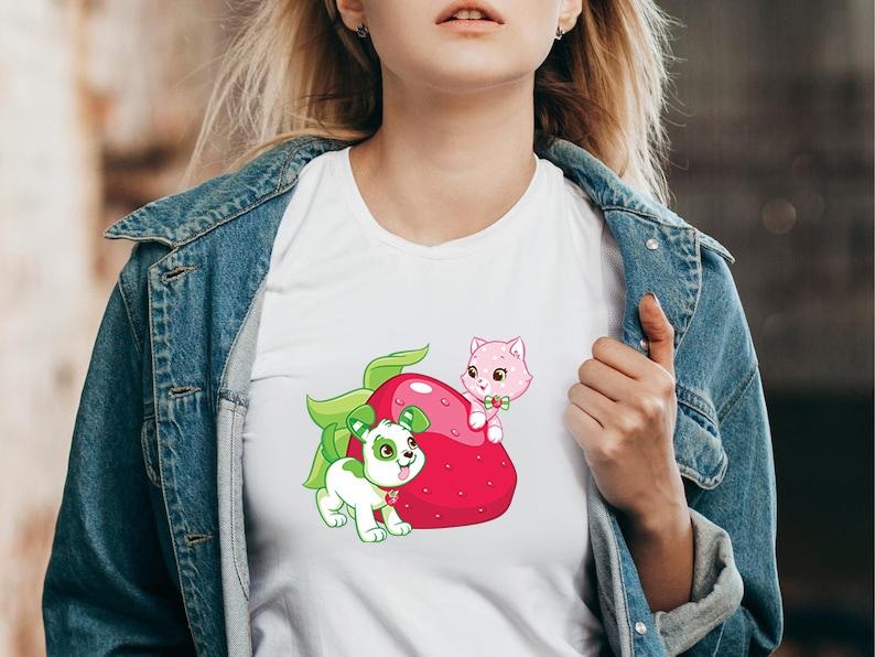May include: White t-shirt featuring a cartoon strawberry with a green leaf, a pink cat, and a green and white dog. The cat is perched on the strawberry, and the dog is standing next to it. The shirt is worn by a person.