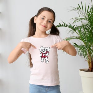 Pu&ograve; includere: T-shirt rosa chiaro con una mascotte di elefante dei cartoni animati che indossa una maglia rossa con una "A" bianca sul davanti. L'elefante &egrave; in piedi con le braccia lungo i fianchi. La maglietta &egrave; indicata da una persona.