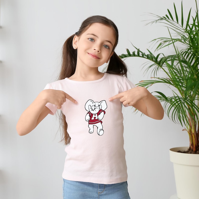 Pu&ograve; includere: T-shirt rosa chiaro con una mascotte di elefante dei cartoni animati che indossa una maglia rossa con una "A" bianca sul davanti. L'elefante &egrave; in piedi con le braccia lungo i fianchi. La maglietta &egrave; indicata da una persona.