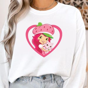 May include: White sweatshirt featuring a cartoon graphic of a girl with pink hair and a strawberry hat, holding a pink cat inside a pink heart. The graphic is centered on the front of the long-sleeved top.