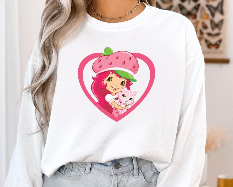 May include: White sweatshirt featuring a cartoon graphic of a girl with pink hair and a strawberry hat, holding a pink cat inside a pink heart. The graphic is centered on the front of the long-sleeved top.