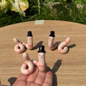 May include: Three small, light pink ceramic worm figurines, each featuring a smooth, curved body with a darker pink band and a tiny black top hat. Each worm has a simple, hand-painted face with two small black dots for eyes and a curved line for a mouth.